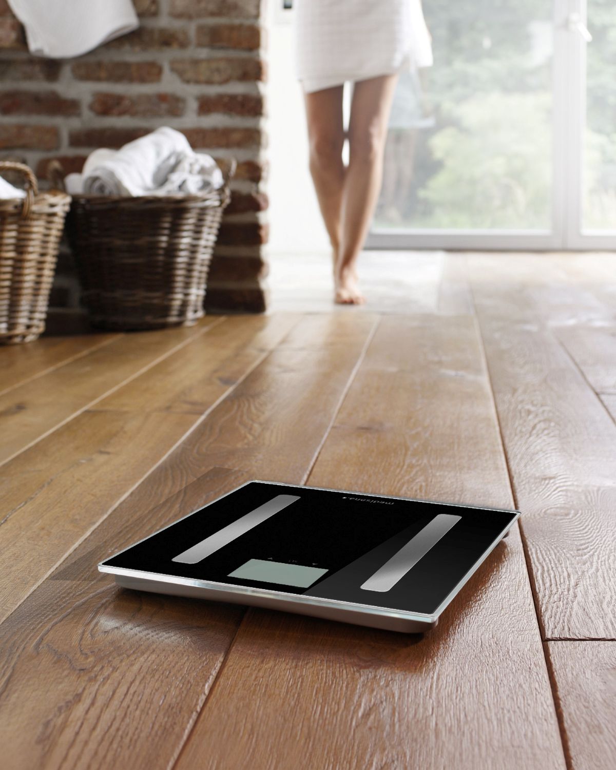 Eine schwarze Körperanalysewaage steht auf einem Holzboden im Badezimmer, während eine Person im Hintergrund auf sie zugeht.